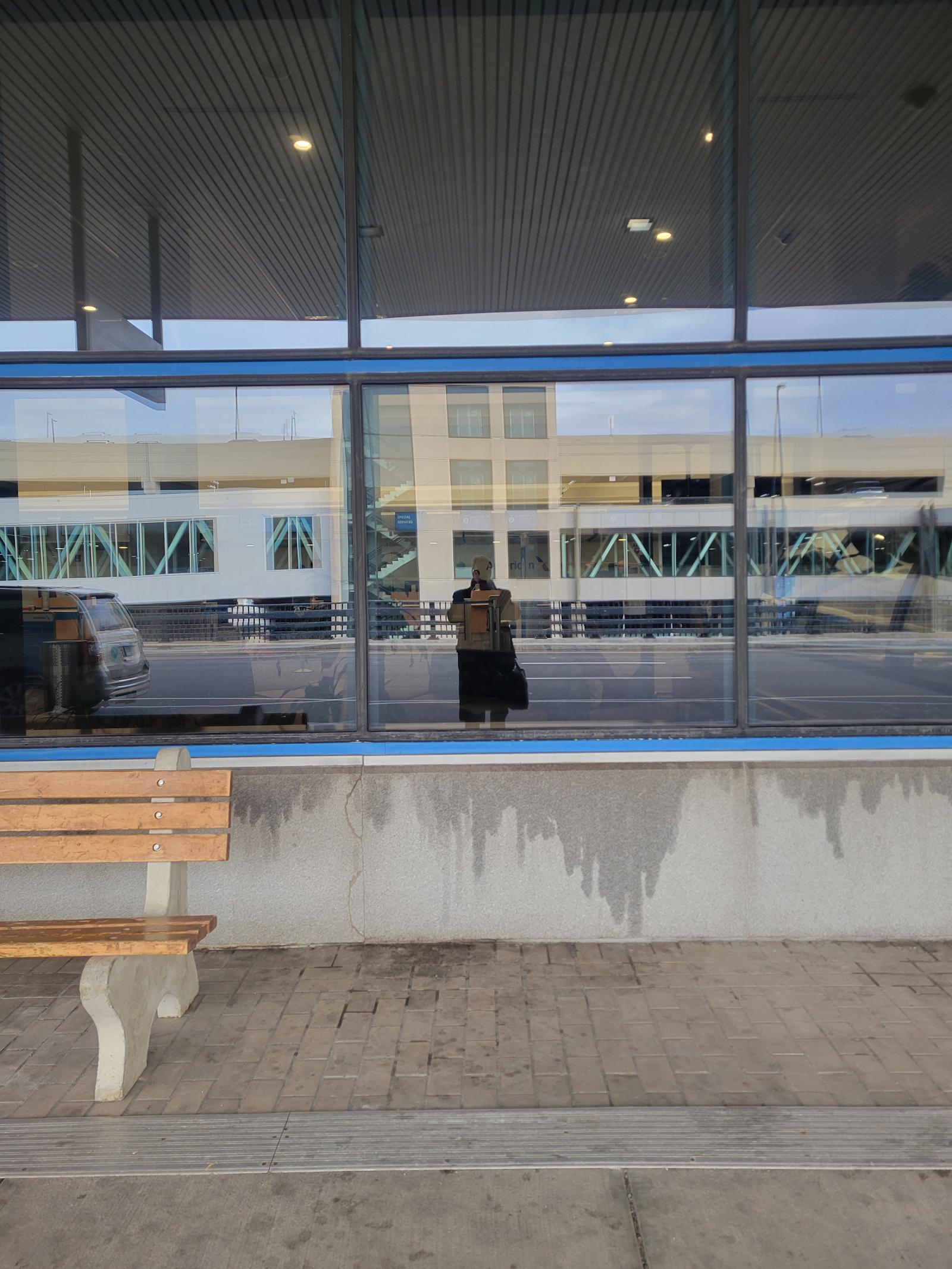 Artist's reflection visible through airport terminal glass from outside, empty bench in foreground, Bradley International Airport March 30 2026 - closing image of What Water Remembers